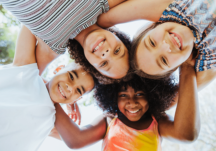 Group of 4 children smiling with arms locked down at a camera on a summer afternoon
